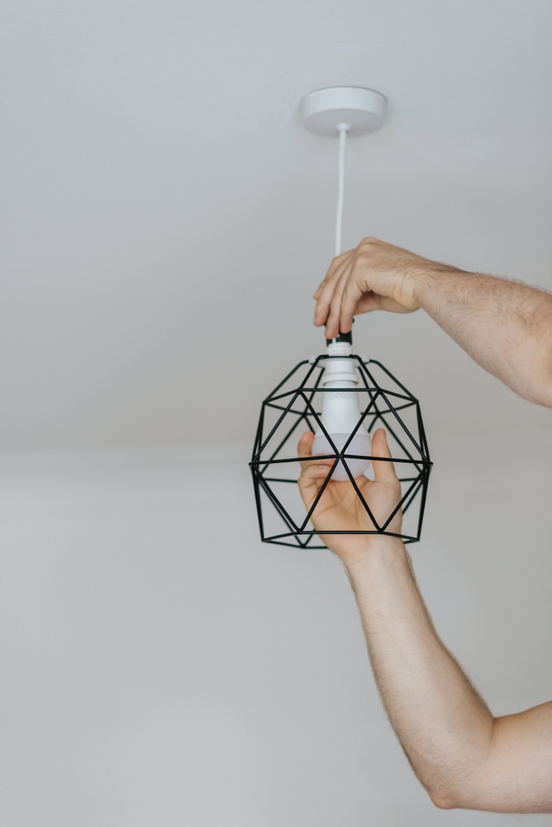 Crop anonymous male screwing bulb into metal lantern hanging on ceiling in house with white wall